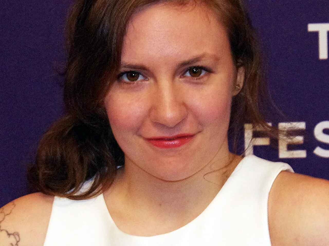 Lena Dunham i vit klänning framför en lila pressvägg på Tribeca Film Festival 2012